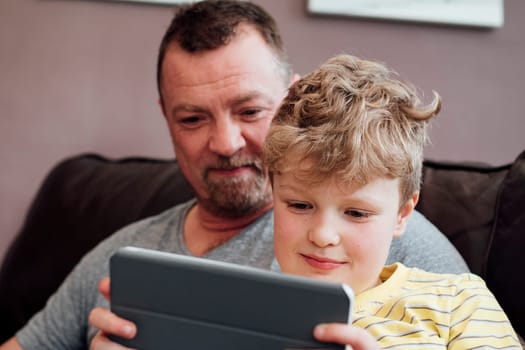 Little boy is sitting on his dads knee on the sofa at home, using a digital tablet. His dad is watching him.