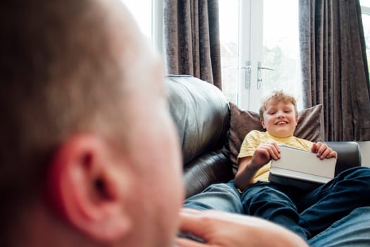 Little boy and his father are talking while lying on the sofa together. The little boy is holding a digital tablet.