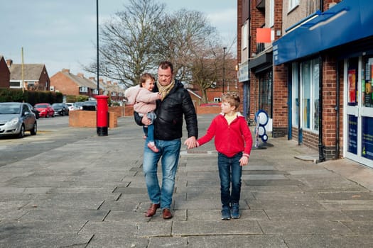 Mature man is taking his children to the local shop with him. He is holding his sons hand and carrying his baby daughter on his hip.