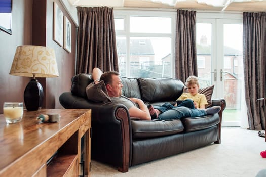 Little boy and his father are relaxing atr home at the weekend. They are sitting at either end of the sofa and the little boy is using a digital tablet while his father watches television.