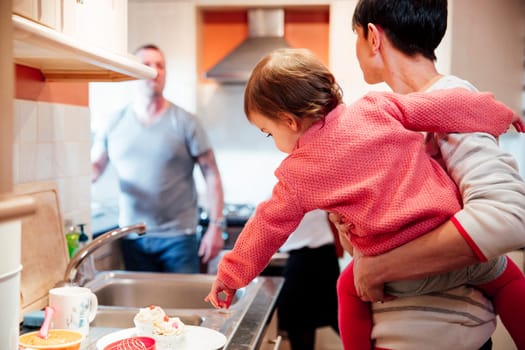 Hungry baby wants the cupcakes that she has spotted on the kitchen worktop. She is on her mother's hip, pointing to the cakes but her parents are busy talking to eachother.
