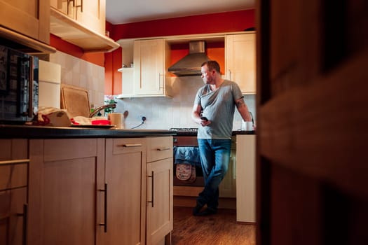 Mature man is standing in the kitchen of his home with a cup of tea in his hand, staring deeply out of the window.