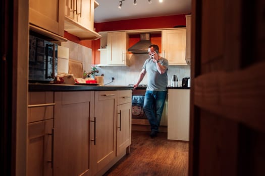 Mature man is standing in the kitchen of his home, looking out the window while talking to someone on his smartphone.