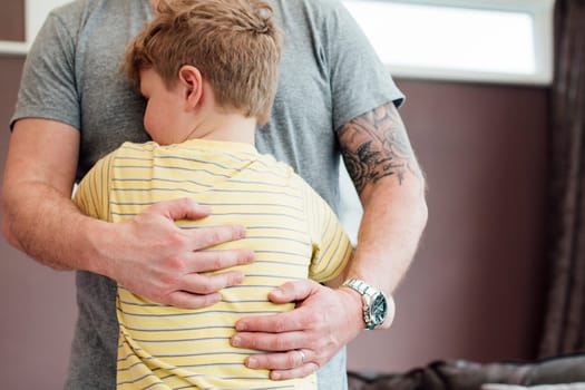 Little boy is cuddling his father at home. He has his arms round his fathers torso and the father is holding his son close to him.