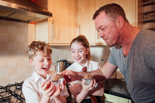 Two children are in the kitchen of their home after school. theyare eating cupcakes and the father is stealing some icing from the top of his sons cake.