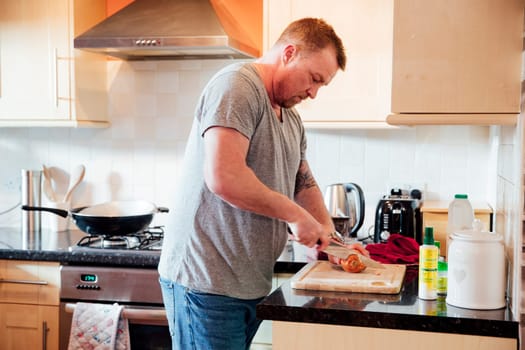 Mature man is cooking in the kitchen of his home. He is cutting up an onion on a chopping board.