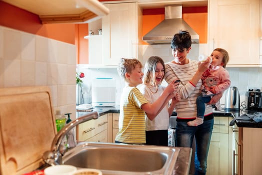 Girl and her brother are showing their mum something on their smartphone. They are all laughing and the mother has the baby on her hip, feeding her at the same time.