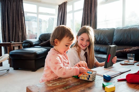 Little girl is distracting her older sister while she is trying to study at home. They are playing with toy blocks together.