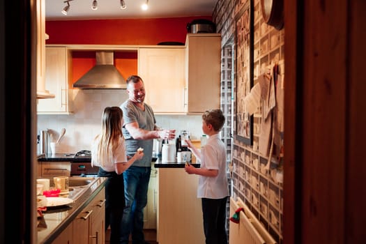 Mature man is with his children in the kitchen of their home getting some snacks. The little boy is taking a glass of milk from his father and the little girl is eating biscuits.