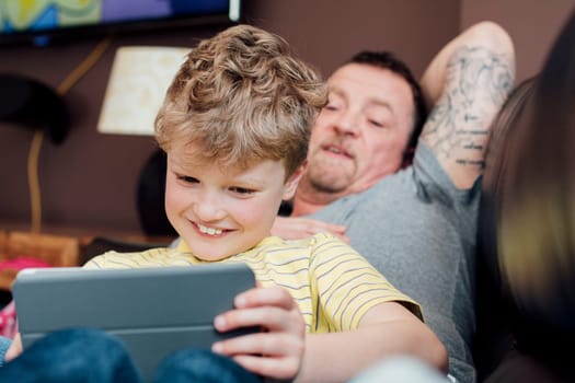 Little boy is sitting on his dads knee on the sofa at home, using a digital tablet. His dad is watching him.