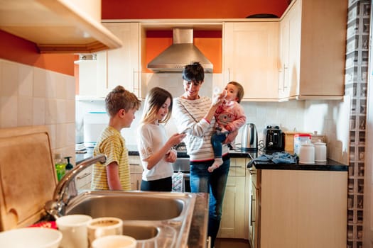Multitasking mum is tending to all of her children at once in the kitchen of their home. She is feeding the baby a bottle while talking to her eldest children and looking at their smart phone.