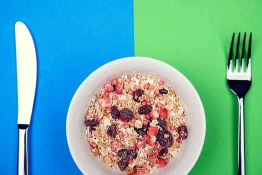 muesli in white bowl on blue and green ground with knife and  fork from above