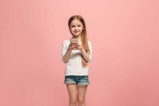 Happy teen girl standing, smiling with mobile phone over trendy pink studio background. Beautiful female half-length portrait. Human emotions, facial expression concept.