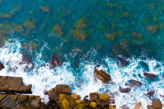 Top aerial view of blue waves crashing on rocky Australian coastline. Summer seascape with birds eye view shot over ocean waves and cliffs. Travel concept background
