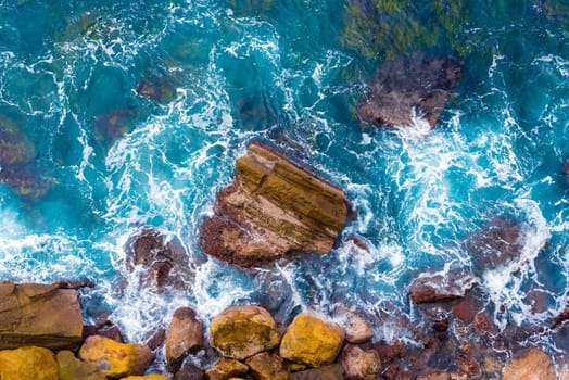 Top aerial view of blue waves crashing on rocky Australian coastline. Summer seascape with birds eye view shot over ocean waves and cliffs. Travel concept background