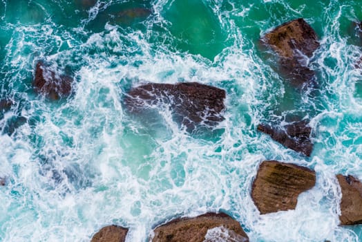 Top aerial view of blue waves crashing on rocky Australian coastline. Summer seascape with birds eye view shot over ocean waves and cliffs. Travel concept background
