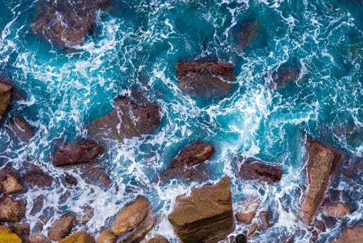Top aerial view of blue waves crashing on rocky Australian coastline. Summer seascape with birds eye view shot over ocean waves and cliffs. Travel concept background