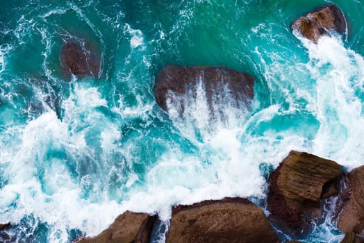 Top aerial view of blue waves crashing on rocky Australian coastline. Summer seascape with birds eye view shot over ocean waves and cliffs. Travel concept background