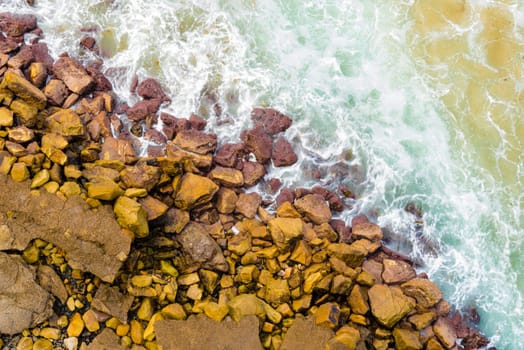Top aerial view of blue waves crashing on rocky Australian coastline. Summer seascape with birds eye view shot over ocean waves and cliffs. Travel concept background