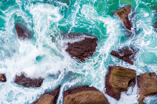 Top aerial view of blue waves crashing on rocky Australian coastline. Summer seascape with birds eye view shot over ocean waves and cliffs. Travel concept background