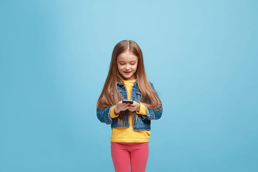 Happy teen girl standing, smiling with mobile phone over trendy blue studio background. Beautiful female half-length portrait. Human emotions, facial expression concept.