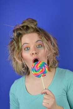 portrait of a young blond woman with colourful streaks in the hair and a big sweet multicolored lollipop in her hand looking surprised into the camera