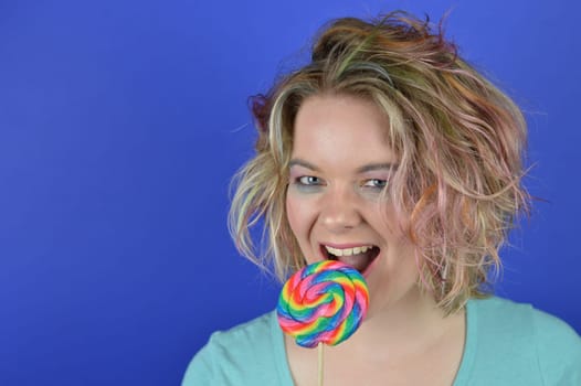 portrait of a young blond woman with colourful streaks in the hair biting into a big sweet multicolored lollipop and having fun looking into the camera