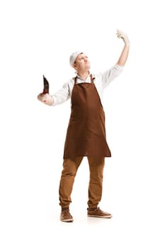 Smiling butcher posing with a knife and mobile phone making selfie photo isolated on white studio background. The young caucasian male model in full-length. Butcher, knife, professional concept