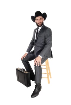 A young handsome business man wearing a suit and cowboy hat sitting.on a bar chair with his briefcase, isolated for white background.