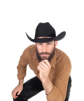 A young man sitting on a chair bending forwards wearing a black.cowboy hat and one hand on his chin, isolated for white background.