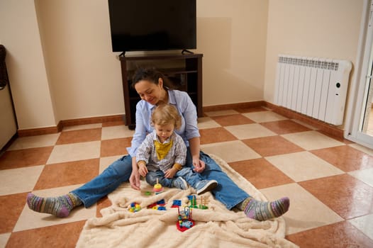 A mother and child enjoy quality time playing together on a blanket indoors. Bright toys and a cozy atmosphere portray care, happiness, and bonding.