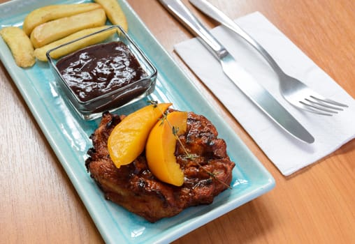 Grilled pork steak fillet with Peach and French fries on blue plate and cutlery over wooden table