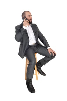 A handsome young business man sitting on a bar chair talking on his.cell phone, with a beard and suit, isolated for white background.