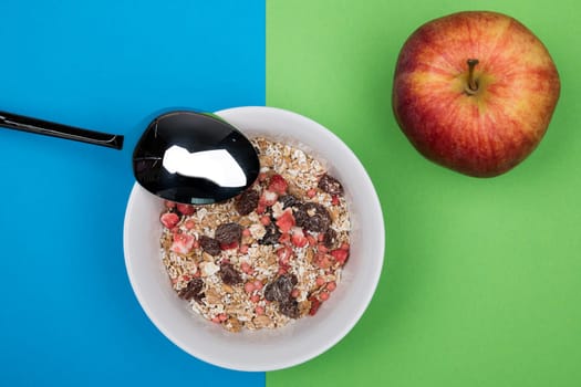 muesli in white bowl on blue and green ground with spoon and apple from above