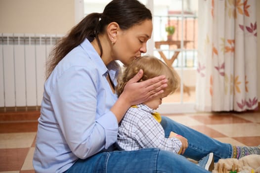 A caring mother kisses her child while sitting together in a cozy, sunlit room, expressing warmth, love, and emotion.