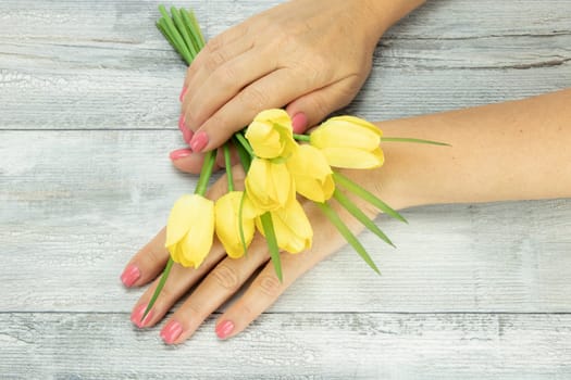 Female hands of woman pink manicured on nails with a bouquet of yellow tulips. Background is a rustic bright table. Concept beauty.