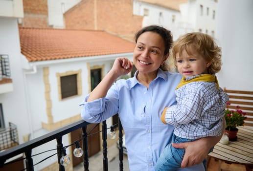 A cheerful mother happily holds her child on a sunny balcony, symbolizing maternal love, family care, and joyful emotions.