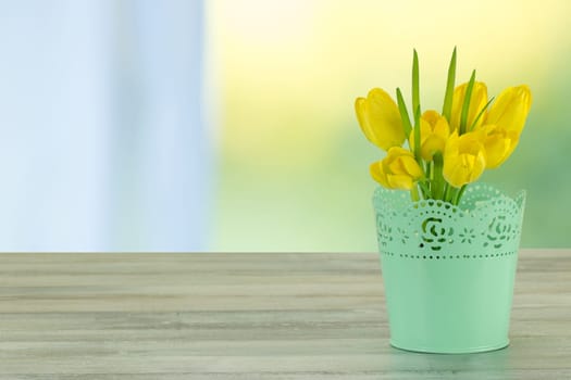 Decorative composition of yellow tulips on rustic bright wooden table. The shining bright background is symbol of spring. The bunch of yellow tulips is in decorative green vase. Empty space for your design. Card concept for mothers day, birthday, aniversary and other events.