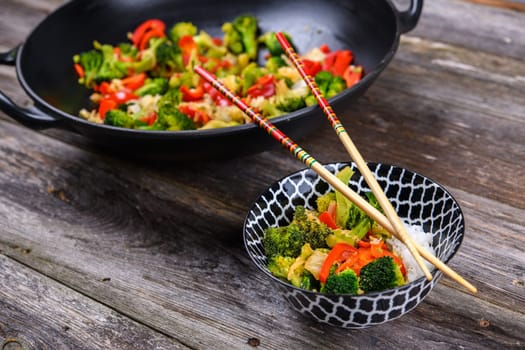 wok with colorful vegetables, bowl of rice and chopsticks on wooden ground