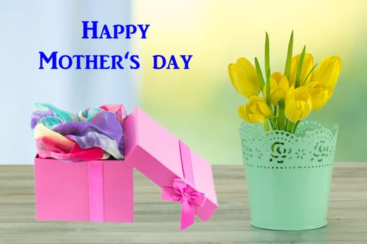 Mothers day greeting card. Decorative composition of yellow tulips on rustic bright wooden table and gift box. The bunch of yellow tulips is in decorative green vase.