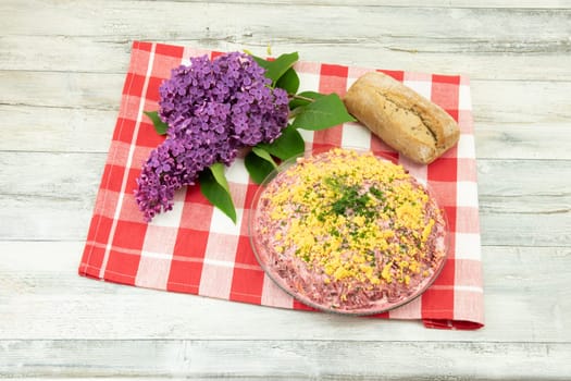 Traditional russian layered salad named herring under a fur coat. Traditional Russian cusine presented on a rustic wooden table at red checkered tablecloth. Next to it a fresh purple lilac blossom and a bread. Concept Russian food.