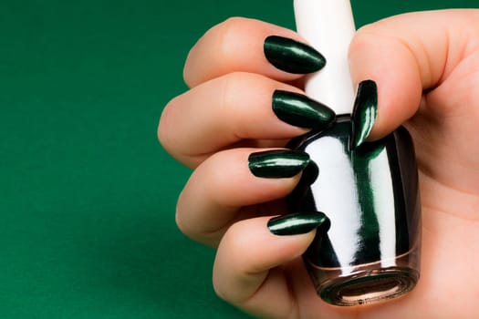 Female hand with dark green nails is holding dark green nail polish on green background. Manicure and nail care concept.