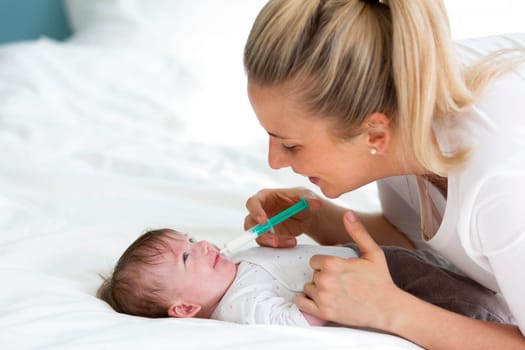 beautiful blond woman and mother, or doctor is feeding baby medicine with a medical syringe on a bed