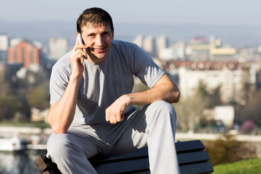The smiling man is using phone and sitting on the bench outdoors.