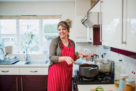 Mature woman is talking and laughing with someone out of the frame while she makes dinner at the cooker.