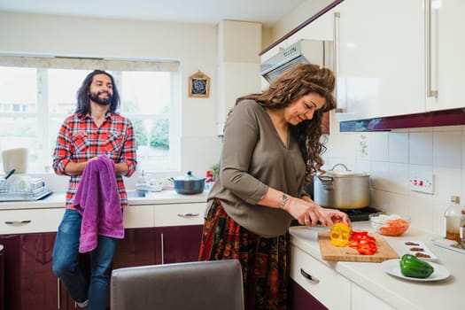 Mid adult man is relaxing in the kitchen talking to his mother while she prepares a curry on the cooker.