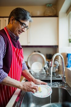 Mature man is doing the dishes in the kitchen of his home. He is wearing an apron while washing a small plate at the sink.