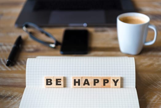 Closeup on notebook over wood table background, focus on wooden blocks with letters making BE HAPPY words. Business concept image. Laptop, glasses, pen and mobile phone in a defocused background.