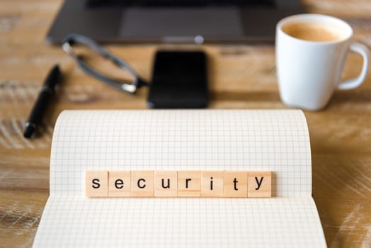 Closeup on notebook over wood table background, focus on wooden blocks with letters making Security word. Business concept image. Laptop, glasses, pen and mobile phone in a defocused background.