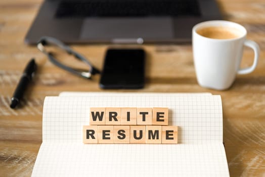 Closeup on notebook over wood table background, focus on wooden blocks with letters making WRITE RESUME words. Business concept image. Laptop, glasses, pen and mobile phone in a defocused background.